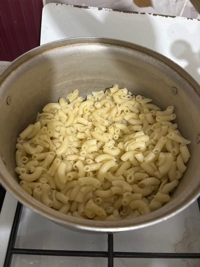 A pot of cooked elbow macaroni is shown from a top-down perspective. The pasta is a pale yellow color and appears to be slightly glistening, suggesting it may have been recently drained. The macaroni is piled high in a silver-colored metal pot. The pot itself is placed on a black stovetop, with some of the stovetop's surface visible at the bottom of the image. The background is out of focus but appears to be a kitchen environment with a white countertop and some red paneling. There are no people visible in the image, and no specific indication of the city of Княгинино, Russia.