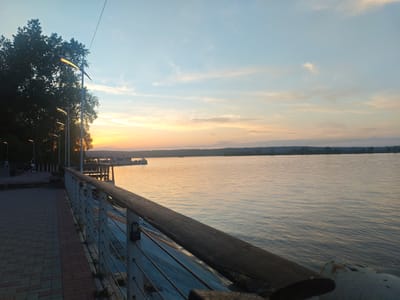 The image shows a sunset over a body of water from a promenade in Krasny Klyuch, Russia. A wooden handrail runs along the right side of the image, supported by metal posts with horizontal bars. A brick-paved walkway is visible to the left of the handrail, with trees and lampposts along the edge of the walkway. In the background, a body of water reflects the orange and yellow colors of the sunset. There is a pier and a boat visible on the water, and land is visible on the horizon. The sky is a mix of blue, orange, and yellow. There are a few people seated on benches at the beginning of the promenade.