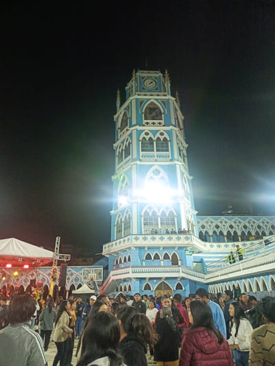 The image shows a nighttime scene in El Cisne, Ecuador, featuring a tall, light blue and white tower, likely part of a church or religious building. The tower has a clock on its upper section and multiple levels with arched windows. Bright lights illuminate the tower. In front of the tower, there is a crowd of people, mainly facing away from the camera. To the left, there is a stage with a white tent-like structure and red stage lighting. People are present on the tower's balconies and walkways. The sky is dark.