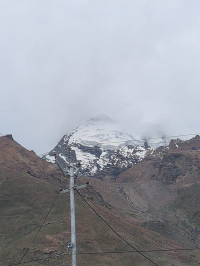 A snow-capped mountain looms in the background under a cloudy sky. The mountain has steep, rocky slopes with patches of snow and ice. In the foreground, a utility pole stands with several wires attached to it, spanning across the image. The terrain around the pole is rocky and barren, with some sparse vegetation visible. The overall impression is of a remote, mountainous landscape. This scene is from Padum, India, a town in the Zanskar region known for its stunning natural beauty and challenging terrain.