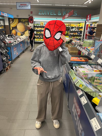 A person with a Spider-Man pillow over their head stands in an Aldi supermarket aisle in Breda, Netherlands. The person wears a grey hoodie and taupe pants with white sneakers. The aisle has shelves stocked with various goods, including toys and food items. A man is visible further down the aisle, examining products. Signs with Dutch text are displayed above the shelves.