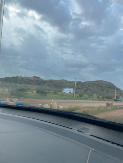 The image depicts a cloudy sky over a hilly, rocky landscape, possibly a rural area near Barakin Columbai, Nigeria. In the foreground, a portion of a vehicle's dashboard and windshield is visible. Beyond the road, there are sandbags and blue barrels, suggesting construction or roadwork. A billboard with several portraits is placed on a pole in the middle ground, against the backdrop of the hills. The overall scene appears to be captured from inside a moving vehicle, given the slight blur and the perspective.