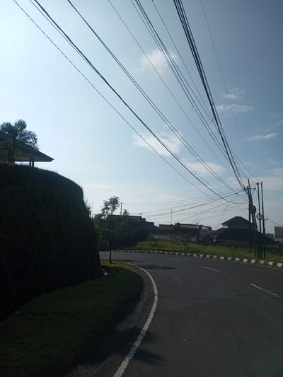 A view of a road with a grassy hill on one side, buildings and power lines in the background under a blue sky with some clouds. This appears to be on the outskirts of a residential area, indicated by the utility poles and power lines running above. The road curves to the right. Based on the location provided, Phouviang-Gnai, Laos, it is likely an arterial road of the municipality with mixed urban and rural features. No people are present in the image.