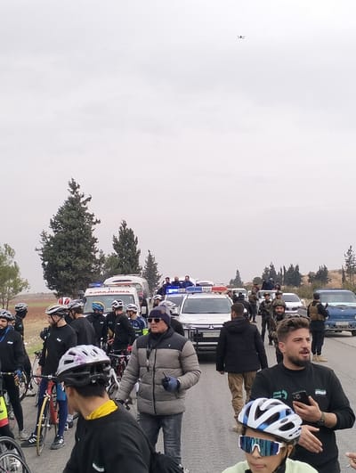 A group of cyclists in black jerseys with helmets are gathered on a road with several support vehicles including police cars and an ambulance. Armed security personnel are visible. One man stands out in a grey puffer jacket. A drone flies overhead. The scene conveys a sense of an organized cycling event with safety and security measures in place. The weather appears overcast.