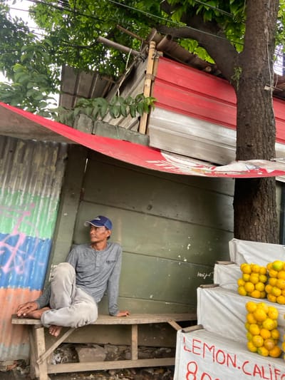 A man in a blue baseball cap and a gray long-sleeved shirt sits on a wooden bench against a green wall. He is looking to the right of the frame. The bench is located in front of a corrugated metal wall painted with blue, green, and pink graffiti. A large, red awning with white stripes extends from the wall. There are stacks of yellow citrus fruit on a white stand to the right of the man. The stand is adorned with a sign that reads "LEMON.CALIF." The scene is situated in the neighborhood of Kecamatan Cimahi Selatan, Indonesia. The location is confirmed by the provided coordinates (latitude: -6.903, longitude: 107.564). A large tree with a thick trunk casts its shadow over the scene.