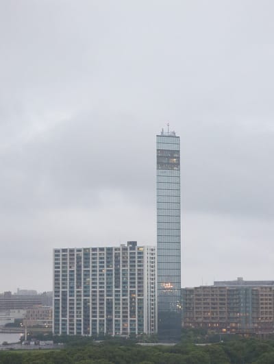 This is a medium-distance shot of a city skyline under a cloudy sky. In the foreground, a dense green tree line runs horizontally across the bottom of the frame. Behind the trees, several buildings are visible. The most prominent is a very tall, slender glass skyscraper with a grid pattern of windows, located slightly to the right of the center of the frame. To the left of this skyscraper is a large, rectangular, light-colored building with many floors and rows of windows, appearing to be an apartment complex. Further to the right of the tall skyscraper, a long, multi-story building with brown-toned balconies and windows is visible. In the background, several other buildings and structures can be seen, hinting at a developed urban area. The overall atmosphere is somewhat muted due to the overcast conditions. This scene is located in Chiba, Japan, likely depicting a modern urban development near the waterfront.