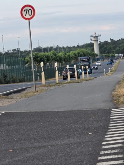 The image captures a scene on a road in Kelsterbach, Germany, likely near the airport given the presence of a control tower-like structure. A speed limit sign indicating "70" is prominently displayed on the left. The road is flanked by trees and a fence. Several vehicles are visible on the road, including trucks and cars. In the foreground, a pedestrian crossing is marked with white stripes on the asphalt. The sky is overcast, suggesting a cloudy day.