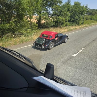 A black classic convertible car with a red soft top is driving on a paved road. The car has a luggage rack on the back with a green cover. The license plate reads "ALC 467A". The driver, a person with fair skin and dark hair, is visible behind the steering wheel. The car is surrounded by green trees and foliage, with a clear blue sky above. The perspective suggests the photo was taken from inside another vehicle. The scene is likely in Naseby, United Kingdom, as indicated.