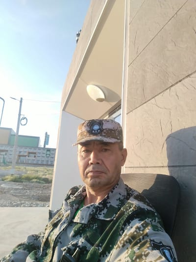 The image shows a man in a camouflage uniform and cap, sitting in a chair outdoors. He is looking directly at the camera with a neutral expression. The background features a modern building with a textured stone facade and a white awning. In the distance, there are several multi-story buildings and some sparse vegetation. A lamppost is visible to the left. The sun is low in the sky, casting long shadows and suggesting it is either early morning or late afternoon. The sky is clear and blue. The man's cap has a patch with text and a globe logo, which appears to be in a non-Latin script. The uniform also has a patch on the sleeve with a similar logo and text. The scene suggests a contemporary, possibly semi-arid, environment.
