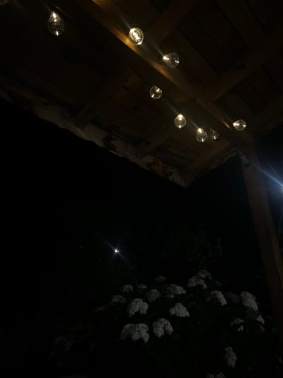 A string of illuminated Edison-style bulbs hangs from a wooden pergola against a dark night sky. The bulbs cast a warm glow on the wooden planks of the pergola's ceiling. Below the pergola, a cluster of white hydrangeas is visible, their blooms faintly illuminated by the ambient light. A bright light source, possibly a streetlamp or distant light, creates a lens flare effect in the upper right portion of the frame. The scene appears to be an outdoor patio or garden area. The location is identified as Salmanovo, Bulgaria.