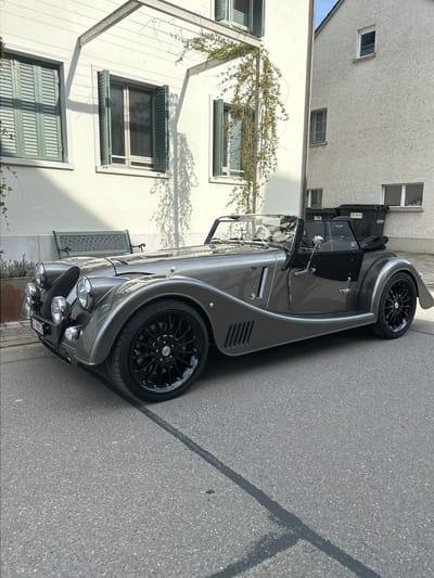 A gray Morgan sports car is parked on a grey asphalt street in Rafz, Switzerland. The car's front is facing the viewer. The license plate is visible. A light-colored building with green-shuttered windows is on the left side of the frame, and a cream-colored building is partially visible in the background. Shadows from foliage are cast on the street and building.