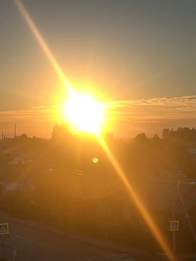 The image captures a brilliant sunset over a residential area, likely in Mezhdurechenskiy, Russia. The sun, a powerful orb of golden light, dominates the sky, casting long, dramatic rays across the scene. The light flares intensely, obscuring many details and creating a warm, hazy atmosphere. In the foreground, a road is visible with a pedestrian crossing sign, indicating a populated area. Beyond the road, a cluster of houses with corrugated roofs are nestled among trees. In the distance, the silhouette of a tall industrial tower or communication mast pierces the horizon, a stark contrast to the natural beauty of the sunset. The sky transitions from a pale blue at the top to a warm orange hue near the horizon, with faint, wispy clouds catching the fading sunlight. The overall impression is one of a serene, yet powerful, natural phenomenon unfolding over a quiet town.