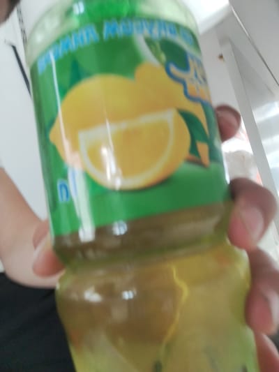 A hand holds a green plastic bottle filled with a yellow liquid. The bottle has a label with the image of lemons and green leaves. The label has text in blue and yellow. The bottle is tilted and partially obscured by a person's hand. The background is blurry and appears to be indoors, possibly a kitchen or a shop. The location is not identifiable from the image itself.