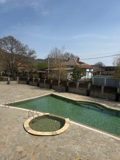 The image depicts a swimming pool area in Sadievo, Bulgaria. The pool has a long rectangular shape with a circular section at one end. The pool's water is clear and slightly green. The pool is surrounded by a stone-tiled area, and there are metal ladders for entering and exiting the pool. Buildings and trees can be seen in the background, along with the sky above. The houses look like a summer vacation spot with stone fences and balconies. The sky is clear. The photograph is taken from an elevated viewpoint.