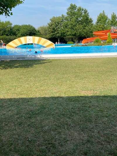 The image depicts an outdoor swimming pool area in Deutschkreutz, Austria. In the foreground, a well-maintained lawn is visible, with shadows cast by unseen trees. The middle ground features a bright blue swimming pool with several people swimming and wading. An arched structure, yellow and white striped with a central logo, spans part of the pool. In the background, vibrant green trees form a dense backdrop. To the right, a large orange waterslide complex is prominent. The sky above is clear and blue.