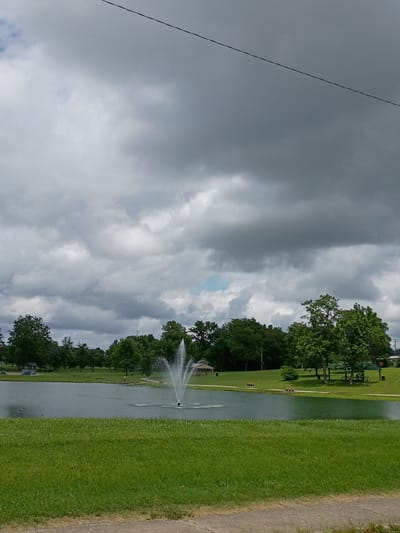 The image shows a serene park scene with a large pond and a fountain in the center. The pond is surrounded by lush green grass and mature trees. In the background, there are more trees and a few scattered structures, possibly gazebos or picnic shelters, suggesting a public park area. A wire stretches across the top of the frame, indicating an overhead utility line. The sky is overcast with dark, heavy clouds, suggesting impending rain or a stormy day. There are no people clearly visible in the image, though a small figure might be present near the pond's edge. The location is identified as Nicholasville, United States of America.