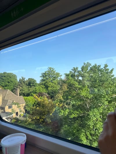 The image, taken from inside a moving vehicle, possibly a train, presents a view of the outside. A portion of a clear blue sky is visible at the left of the frame, bisected by thin, white contrails from an aircraft. Adjacent to the sky, there is a dense arrangement of trees with varied green foliage.

Through the trees, a building with a steeply pitched roof made of stone tiles can be partially seen. The house has small windows and a chimney stack.

The photo is framed by the window of the vehicle. In the lower right corner, a white and pink plastic container is partially visible.