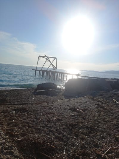 This image captures a scene at the coast of Gwandra, Abkhazia, under a bright, sunlit sky. In the foreground, a pebble-strewn beach is visible, with scattered debris such as twigs and plastic bottles. A large, weathered concrete structure dominates the right side of the mid-ground, providing a stark contrast to the natural elements.
The sea stretches out into the distance, its surface shimmering with sunlight. On the left side of the mid-ground, an old, dilapidated pier extends into the water, its wooden pilings partially submerged. The pier's structure appears damaged and incomplete, with a metal frame on top that is twisted and broken. In the distance, a faint outline of hills or mountains can be seen along the horizon, meeting the calm sea. The sun is prominently positioned in the upper right quadrant of the image, casting a bright glare that partially obscures details in that area. A few silhouetted figures can be observed standing on the pier, appearing as small dark shapes against the bright water and sky. The overall impression is one of a weathered, coastal landscape with remnants of human infrastructure.