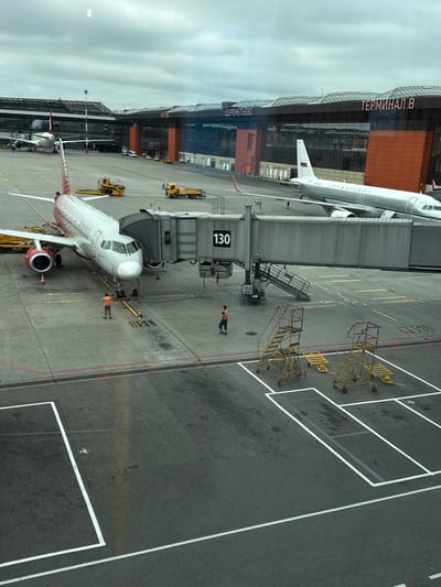 This is an aerial view of an airport tarmac at Sheremetyevo International Airport (SVO) in Rzhavki 2, Russia, specifically Terminal B. Arossia Airlines aircraft is parked at gate 130, connected to the terminal by a jet bridge. Two airport workers in high-visibility vests are on the tarmac near the aircraft. Another Aeroflot aircraft is parked in the background. Several service vehicles, including a tow tractor and a baggage cart, are visible on the tarmac. The sky is overcast.