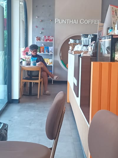 A daytime interior shot shows a man with curly dark hair seated at a table near a window at Punthai Coffee in Plai Phraya, Thailand. He is wearing glasses, a dark shirt, and slides. His legs are crossed and he is looking down at something in his hands, perhaps reading or using a device. A table and chair are in front of him, and a display of packaged goods is visible on a shelf behind him. A counter with brown and orange panels dominates the right side of the frame. In the foreground, a chair with a light brown seat and back is partially visible. The overall lighting is bright and diffused.