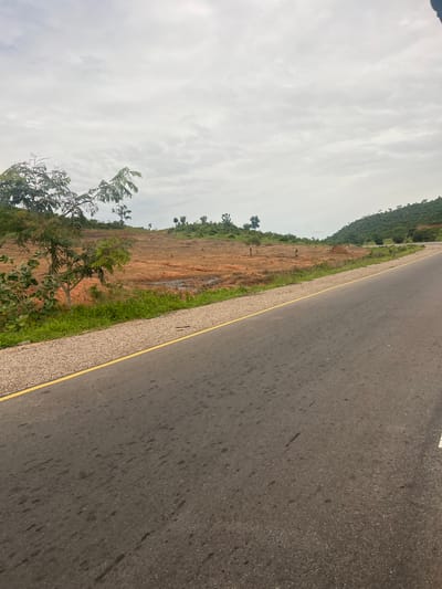 The image depicts a paved road running diagonally through a landscape. The road surface is dark asphalt, with a distinct yellow line marking the edge closest to the viewer. To the left of the road, there is a strip of gravel, followed by a red, earthen hillside dotted with sparse vegetation and some trees. Further to the left, the hillside rises into a densely wooded area, with a green, tree-covered mountain in the background. The sky is overcast with scattered clouds, suggesting a muted light and possibly a day without direct sunlight. The scene appears to be a rural or semi-rural setting in Minda, Nigeria, given the provided location context. There are no people or specific objects that stand out, beyond the natural elements and the road infrastructure. The overall impression is one of a journey or transit through a natural, somewhat undeveloped area.