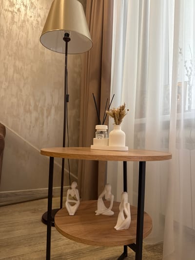 The image shows a room interior. The room features a lamp with a gold-colored shade positioned on the left. To the right is a two-tiered round table. The top tier contains a white vase with dried flowers, a fragrance diffuser bottle, and a sculptural representation of a human figure. The bottom tier also displays human figure sculptures. The background includes a window with white curtains and a wall with textured wallpaper. There are no indications of it being in the city of Goryachy Klyuch, Russia, or any people present.
