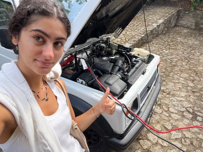 The scene depicts an outdoor setting on a stone-paved surface in Vila Nova de Cerveira, Portugal. A female subject, with dark hair and a light complexion, is positioned in the foreground, capturing a selfie. She is wearing a white ribbed tank top, with a white cable-knit sweater draped over her shoulders. A multi-strand necklace adorned with dark beads and a circular pendant is visible on her chest. Her right hand, positioned in the lower-center of the frame, displays a thumbs-up gesture. Adjacent to the subject, a white utility vehicle, appearing to be an older model SUV or jeep, is present with its hood raised and fully open, exposing the engine compartment. Red and black jumper cables are connected to the vehicle's battery, located within the engine bay, and extend towards the front of the vehicle. The vehicle's front grille is visible. The ground is composed of irregular, light-colored stone pavers. In the background, a low, dry-stacked stone wall is visible, rising from the paved surface. The lighting indicates a bright, clear daytime environment.