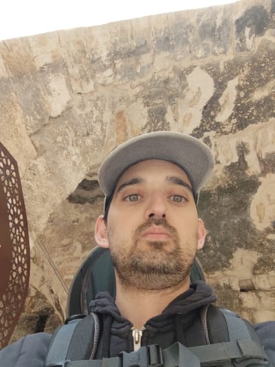 A male subject is centered in a close-up, chest-up shot, looking directly at the camera. He is an adult with a short dark beard and mustache, light-colored eyes, and is wearing a grey baseball cap, a dark grey or black zippered hooded sweatshirt, and a dark grey backpack with visible straps and a black plastic buckle. The background consists of a rough-hewn stone wall, constructed from light-colored blocks, forming an arching structure overhead and behind the subject. A decorative, rusted metal panel with an intricate geometric pattern is partially visible on the left side of the frame. The scene is located within Tel Afek National Park (Antipatris) in Rosh HaAyin, Israel, likely adjacent to the Ottoman fortress ruins.