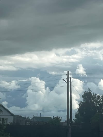 The image depicts a cloudy sky over a rural landscape. In the foreground, there is a dark fence and some trees. Further back, several houses with visible roofs and some chimneys are present, along with power lines supported by a utility pole. The sky is filled with a mix of dark, heavy clouds and lighter, brighter formations, suggesting a potentially stormy or overcast day. The overall scene appears to be a view of a village or small settlement.