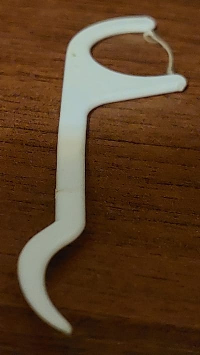 A white dental floss pick with a curved handle and a looped end holding a strand of floss rests on a dark brown, wood-grained surface. The floss appears slightly yellowed or soiled. The object is positioned diagonally across the frame, with the handle curving towards the bottom left. The lighting is overhead, casting soft shadows and highlighting the texture of the wood. There are no people present. The location is not discernible from the image alone.