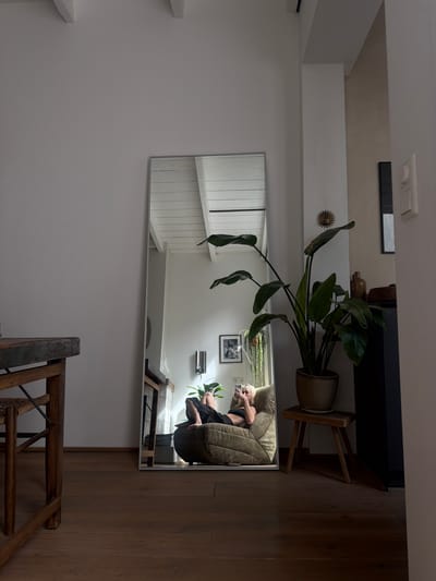 The image depicts an interior scene in a dwelling, possibly a living room, located in Leiden, Netherlands. A full-length mirror dominates the center of the frame, reflecting a woman seated on a plush, olive-green beanbag chair. She is wearing dark athletic wear and holding a smartphone, seemingly taking a selfie. The room is illuminated by natural light streaming from a window or overhead source, casting distinct shadows. To the right of the mirror, a large potted plant with broad leaves stands on a wooden stool. The room features wooden flooring and white-painted walls. In the foreground on the left, a portion of a wooden table with a distressed finish is visible. The background, as reflected in the mirror, shows a white slatted ceiling and a framed picture on the wall.