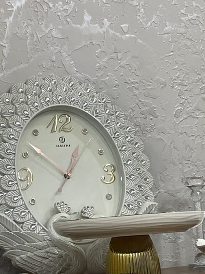 A decorative wall clock with a peacock feather design, featuring a cream-colored face with gold numbers and pink hands, is displayed against a textured gray wall. The clock is embellished with small, clear rhinestones that resemble dewdrops. The clock is supported by a white ceramic swan figure, which is perched on a golden fluted base. A clear glass candlestick holder is visible to the right of the clock. The setting appears to be an indoor environment. This image does not contain any identifiable landmarks or contextual clues that would allow for its location to be determined in the city of Armavir, Armenia.