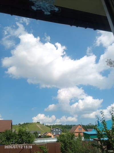 The image shows a view from a window looking out at a rural landscape under a bright blue sky with white cumulus clouds. In the foreground, the dark brown frame of the window is visible at the top and right edges. Below the sky, a line of houses and trees is visible. On the left, a house with a brown roof stands out. Further back, a green, grassy hill with a winding path is prominent, dotted with trees. Several other houses with various roof colors, including blue and orange, are scattered throughout the scene. A greenhouse with a metallic roof is also visible. The bottom edge of the image displays a timestamp: "2026/07/12 15:28". The scene appears to be a residential area in a village or countryside setting, likely in Райманово, Russia, given the context.