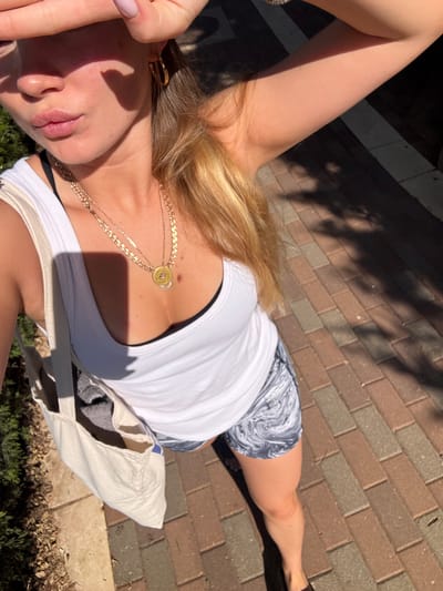 A woman is pictured outdoors in what appears to be Ariel, Palestinian Territories based on the type of paving and vegetation visible. She is lying on a brick-paved surface partially shaded by trees. She is wearing a white tank top, a black sports bra is visible beneath it, and patterned shorts. A gold necklace adorns her neck. She is also carrying a white tote bag with a gray item inside. One of her hands is partially obscuring her face. The environment includes greenery visible in the lower portion of the frame and shade from trees cast on the brick surface.