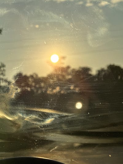 This media file captures a hazy view of the sun through a dirty and scratched surface, likely a window or glass. The sun appears as a bright, glowing orb, casting a warm, diffused light across the scene. In the background, a dark silhouette of trees or foliage is visible, creating a natural setting. The overall atmosphere is softened by the atmospheric conditions and the imperfections on the glass, suggesting a serene, possibly early morning or late afternoon, time of day. The presence of dust and scratches on the surface adds a sense of texture and perhaps a hint of neglect or age. There is no visible text in the image. The location context of Pozhernik, Bulgaria, suggests a rural or natural environment where such a scene might be observed.