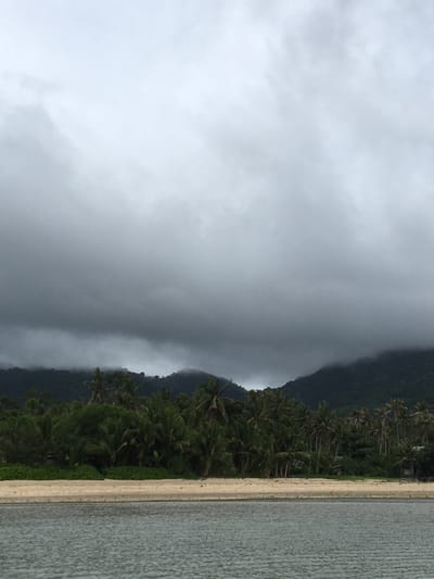 The image depicts a beach scene in Baan Kai, Thailand, on a cloudy day. To the right of the frame is the ocean, next to a tan sandy beach. A line of dense green vegetation, including palm trees, separates the beach from a dark, steep hillside or mountain. Heavy gray clouds cover the sky and obscure the upper portion of the mountain.