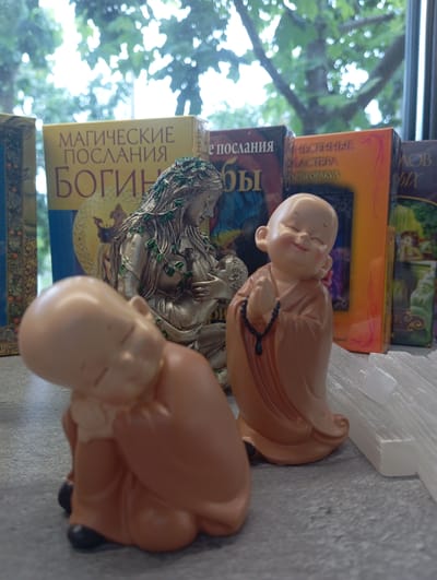 In Tairove, Ukraine, on a gray, textured surface, are several decorative figurines and books. In the foreground, a light-colored figurine of a sleeping baby is positioned to the left, its head bowed and body curled. To the right and slightly behind the baby figurine, a small, smiling monk figurine is depicted with hands clasped in prayer and wearing an orange robe. Behind the figurines, a statue of a woman adorned with green vines and holding a baby is visible. Several books are arranged behind the statues. One book is yellow with Russian text reading "МАГИЧЕСКИЕ ПОСЛАНИЯ" and "БОГИНИ". Another book is orange with Russian text. A third book is dark blue with Russian text, and a fourth book is dark green with an illustration. The background shows blurred green foliage and branches through a window.