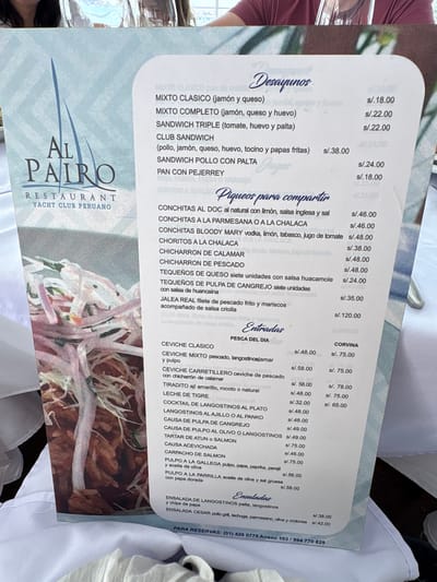 This media file is an image of a menu at the restaurant "Al Pairó Yacht Club Peruano" located in La Punta, Peru. The menu is divided into sections: "Desayunos" (Breakfast), "Piqueos para compartir" (Appetizers to share), "Entradas" (Entrees), and "Ensaladas" (Salads). Each item is listed with its description and price in Peruvian Soles (s/).
The "Desayunos" section offers classic breakfast options like mixed sandwiches with ham and cheese, complete mixed sandwiches with various ingredients, and club sandwiches. The "Piqueos para compartir" section features seafood appetizers such as conchitas (scallops) prepared in different styles like al natural, a la parmesana, or a la chalaca, as well as Bloody Mary conchitas, choritos (mussels), fried calamari, fried fish, and cheese tequeños.
The "Entradas" section includes more substantial seafood dishes like ceviche (classic and mixed), ceviche carretillero (with fried calamari), tiradito, leche de tigre, cocktail de langostinos (shrimp), and various causass (a Peruvian appetizer made with mashed potato). There are also octopus dishes like pulpo al olivo, pulpo a la gallega, and grilled pulpo.
The "Ensaladas" section presents a shrimp salad with avocado and chips, and a Caesar salad with grilled chicken.
The menu also displays the restaurant's name and logo prominently at the bottom left. The contact information for reservations, including a phone number and extension, is visible at the bottom right. The overall presentation is clean and organized, suggesting a professional establishment. The menu appears to be for a lunch or dinner service, given the variety of seafood and main courses. The image itself is taken from a slightly overhead angle, capturing the entire menu. The lighting suggests it's daytime, possibly indoors with ambient light. There are no people clearly visible within the frame of the menu itself, but the context implies a dining setting.