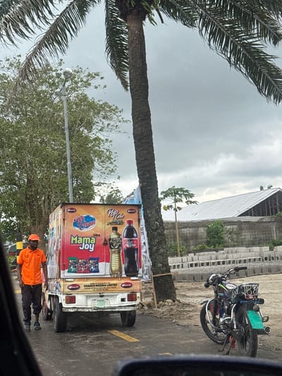 The image depicts a street scene in Igwuruta, Nigeria. A truck displaying advertisements for "Pop Cola," "Too Clean," and "Mama Joy" is parked on the side of the road. A man wearing an orange shirt and cap is standing beside the truck. Palm trees are visible in the background, and the sky is overcast. A motorcycle is parked on the sidewalk. A building is situated next to stacked gray blocks.