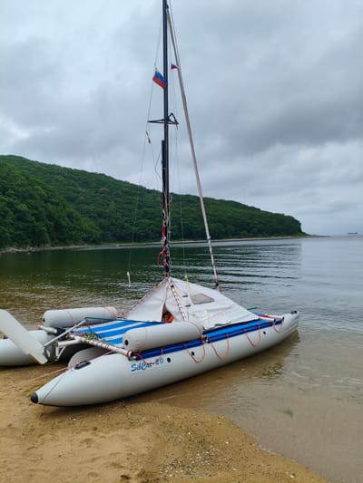 The image shows an inflatable catamaran, identified by the text "SibCat-20" on its side, resting on a sandy shore at the edge of a body of water. The catamaran has two large inflatable hulls and a central deck covered with a white tarp, with blue trim along the edges. A mast stands tall, with rigging and two Russian flags attached. The water is calm and reflects the overcast sky. Behind the water, a lush green, tree-covered hillside rises, characteristic of the landscape around Risovaya Pad'. The sky is cloudy and gray, suggesting a potentially cool or damp day. The scene is serene, with no immediate human activity visible in the foreground, though a few distant figures can be observed on the shore of the bay in the middle ground. The overall impression is one of a quiet, natural coastal setting.