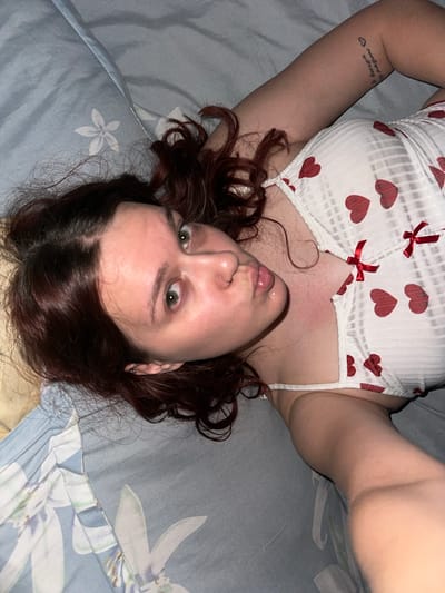 This media file depicts a person lying on their back on a bed with a floral-patterned blue and white comforter. The person has long, dark, curly hair and is wearing a white ribbed tank top with a pattern of red hearts and small red bows. The person's face is visible, looking directly at the camera with wide, green eyes. Their lips are slightly parted. A small tattoo is visible on their left arm. The image appears to have been taken indoors, likely in a bedroom, as suggested by the bed and comforter. The lighting suggests it might be nighttime or a dimly lit room. The overall mood conveyed by the image is intimate and direct, with the subject making eye contact with the viewer. There is no visible text in the image. The location context of Bishkek, Kyrgyzstan, does not provide specific visual cues within the image itself.
