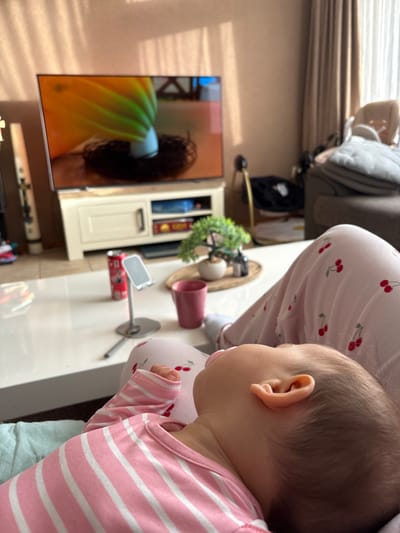 The image shows a baby in a pink and white striped shirt, facing away from the camera, with a pacifier in its mouth. The baby is sitting next to a person wearing cherry-patterned pajamas. In the background, there's a television displaying an image of a person with dark hair and a yellow and green object. Beside the TV, there is a cabinet with decorative items and a plant on top. A red can and a small white object are also on the table next to a pink mug. The setting appears to be a living room interior. The location in the city of Veghel, Netherlands is not evident in the image.