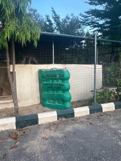 The image shows the entrance to Aldusar Zoo in Batagarawa, Nigeria. A large, green, sculpted object with the text "Welcome to ALDUSAR ZOO FIRST OF ITS KIND IN KATSINA 3 MAY 2019" is visible. Behind the sculpture, a fenced enclosure contains an animal, possibly a deer or antelope, standing on its hind legs. A tree with long, green leaves is in the foreground on the left. The enclosure is topped with a dark roof and surrounded by a fence. A paved area with a black and white striped curb is in the foreground. The sky is blue with some clouds.