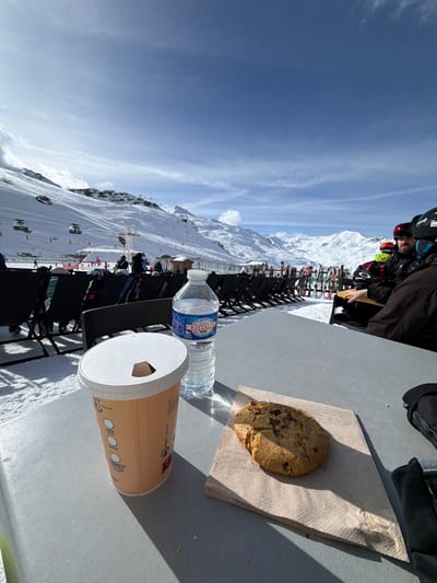 The scene depicts an outdoor rest area on a snow-covered terrace within a ski resort in Les Belleville, France. In the foreground, a light grey table holds a light orange-brown disposable cup with a white lid featuring a hexagonal opening, a transparent plastic bottle of Cristaline mineral water with a blue cap, and a large chocolate chip cookie resting on a light brown paper napkin. A black ski glove is partially visible on the table's right edge. The midground reveals rows of black outdoor chairs, some occupied by individuals dressed in ski attire including helmets and jackets. To the right, multiple pairs of skis and poles are racked vertically in the snow. On the left, a ski lift system with several dark gondola cabins ascends a snow-covered mountain, where other skiers are visible on groomed slopes. The background presents vast snow-covered mountain ranges under a clear, bright blue sky with sparse white clouds. Strong sunlight casts distinct shadows across the foreground elements and the snow.