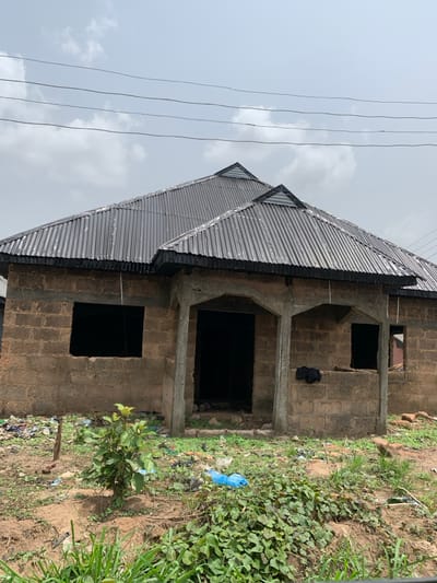 An unfinished, single-story structure with exposed concrete block walls and a dark grey corrugated metal roof stands under an overcast sky in Okokpon, Nigeria. The building features a front facade with two concrete pillars forming a recessed entrance area; one archway leads to a main dark void entrance, and another archway is beside a smaller, open window aperture. A larger rectangular window opening is present on the left side of the facade. The ground surrounding the building is uneven, composed of dirt and sparse green vegetation, including small plants and ground cover. Various pieces of scattered debris and refuse, including plastic bags, are visible on the ground. Multiple parallel utility wires span across the upper portion of the sky above the building. No individuals are present in the image.