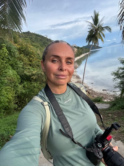 In the foreground, a woman with blonde hair tied back, facing forward, takes a selfie. She is wearing a long-sleeved, light green t-shirt with "USERA SPORT" visible on the left chest. A black camera strap crosses her chest, and a light-colored bag is slung over her right shoulder. She holds a black Sony camera with a lens in her right hand. Her skin appears tanned with a slight sheen.
The background reveals a tropical landscape. Lush green foliage covers a hillside to the left, with a clear blue sky above. To the right, a calm body of water, possibly the sea, meets a sandy shoreline dotted with debris. A prominent palm tree leans over the water, its fronds extending towards the top left of the frame. In the distance, hills rise from the water under a cloudy sky. The scene is consistent with a coastal area in Baan Pang Ka, Thailand.