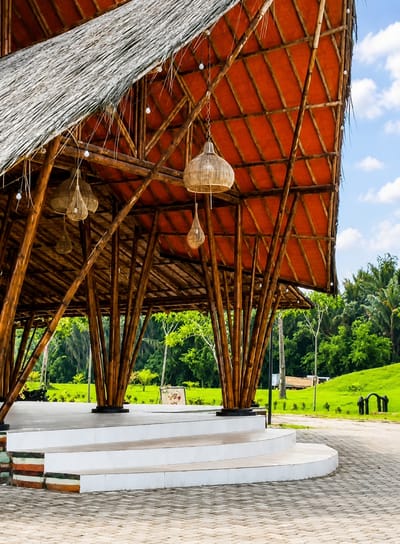 The image depicts an open-air pavilion constructed primarily from bamboo. The roof is thatched with dried grass on the exterior and features a vibrant orange-colored fabric or material on the interior, supported by a complex network of bamboo poles. Several woven pendant lights hang from the ceiling. The pavilion has a raised white stage area with steps in the foreground. Behind the pavilion, a lush green landscape is visible with trees and a grassy hill. A pathway paved with interlocking bricks leads around the stage. The sky is blue with scattered white clouds. There are no people visible in the scene. The location is identified as Eampat Kwalamencirim, Indonesia.