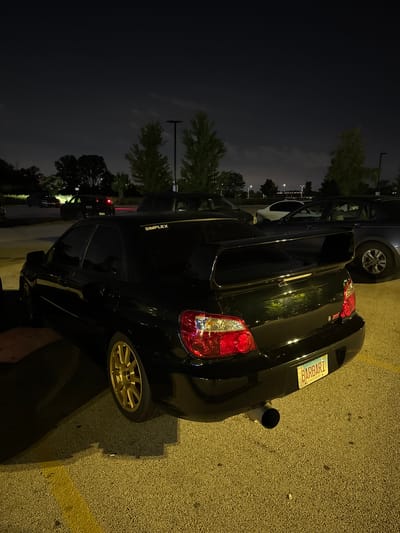 In the city of Schaumburg, United States of America, a black Subaru WRX STI is parked in a dimly lit outdoor parking lot at night. The car, viewed from its rear three-quarter angle, exhibits a large rear wing, gold multi-spoke rims, and a visible exhaust pipe. The license plate reads "BARBARI" and is bordered by a yellowish frame with the words "LAND OF LINCOLN" printed below. Above the rear window, a decal reads "SIMPLEX". The parking lot's asphalt surface is marked with a yellow line. The background features trees and streetlights, suggesting an urban or suburban setting.