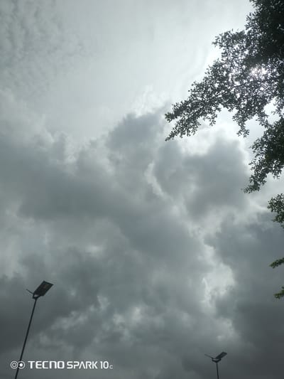 The image, taken in Awoyaya, Nigeria, depicts a sky filled with dense, grey clouds, suggesting an overcast or approaching stormy weather condition. Sunlight filters through a gap in the clouds at the top right of the frame, creating a bright, diffused glow. In the foreground, the silhouette of tree branches with small leaves frames the upper right portion of the sky. Two streetlights are visible: one tall pole with a solar panel light fixture stands in the lower left quadrant, and a second, shorter pole with a similar light fixture is positioned in the lower right corner. The bottom of the image features a watermark that reads "TECNO SPARK 10c". There are no people or animals visible in the scene. The overall environment appears to be an outdoor setting in a developed area, indicated by the presence of the streetlights.
