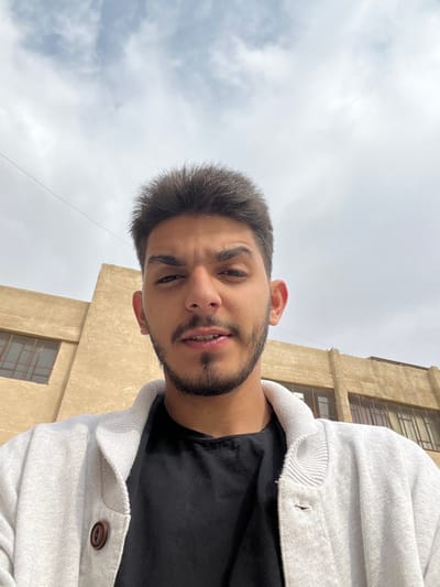 A young man takes a selfie outdoors. He has short dark hair, a beard, and is wearing a black shirt and a light-colored button-down jacket. The sky is overcast and cloudy. Behind him is a tall, beige building with windows. The camera angle is low, looking up at the man.